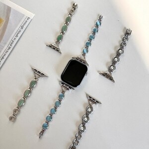May include: A silver smartwatch with a black screen is surrounded by four decorative watch bands. The bands feature green, blue, and white gemstones, and a pearl chain. The bands are designed to be interchangeable.