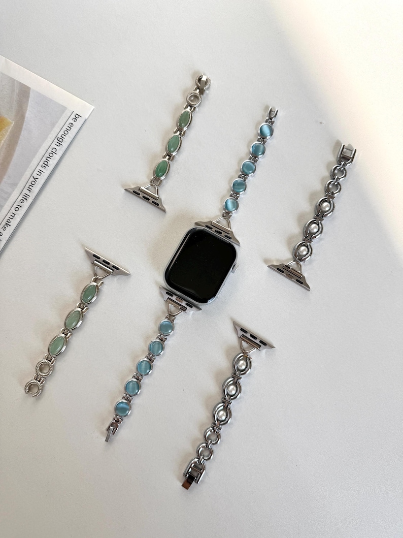 May include: A silver smartwatch with a black screen is surrounded by four decorative watch bands. The bands feature green, blue, and white gemstones, and a pearl chain. The bands are designed to be interchangeable.