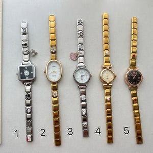 Puede incluir: Cinco relojes de mujer con diferentes diseños. Los relojes tienen correas doradas y plateadas, con varias formas de esfera y adornos. Un reloj tiene una esfera cuadrada negra, mientras que otro tiene una esfera ovalada dorada. Los relojes se muestran sobre una superficie clara.