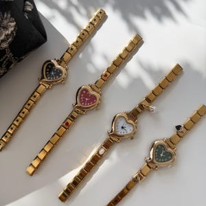 May include: Four gold-toned wristwatches with heart-shaped faces. Each watch has a different colored face: black, red, white, and green. The watch bands are rectangular links. A black floral fabric is in the background.
