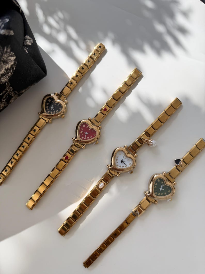 May include: Four gold-toned wristwatches with heart-shaped faces. Each watch has a different colored face: black, red, white, and green. The watch bands are rectangular links. A black floral fabric is in the background.