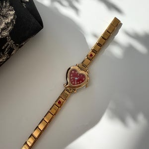 May include: Gold-toned wristwatch with a heart-shaped red face. The watch has a gold band with small heart accents. The watch is on a white surface, with a black patterned pouch in the background.