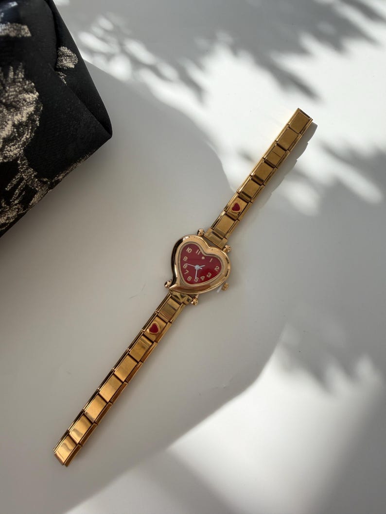 May include: Gold-toned wristwatch with a heart-shaped red face. The watch has a gold band with small heart accents. The watch is on a white surface, with a black patterned pouch in the background.