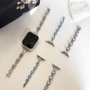 May include: Silver-toned watch bands with various gemstone and pearl accents. One band is attached to a black smartwatch, while others are detached. The bands feature oval green, blue, and white gemstones, and pearls. The bands are designed for a smartwatch.