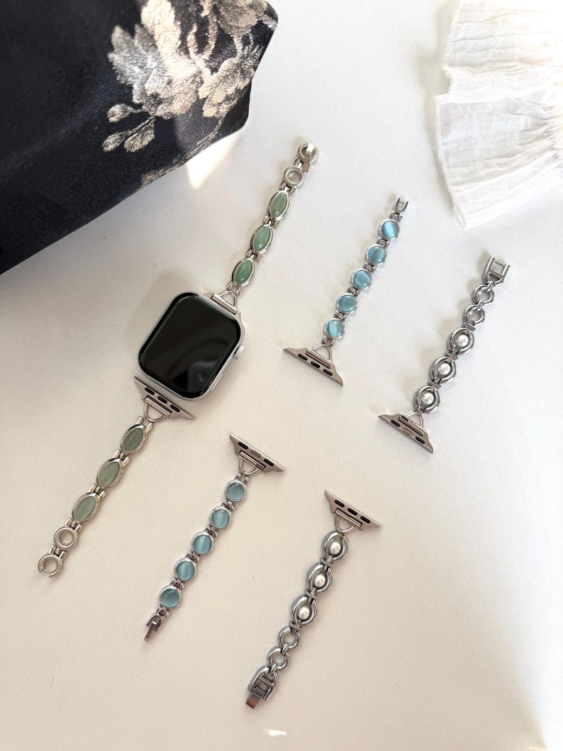 May include: Silver-toned watch bands with various gemstone and pearl accents. One band is attached to a black smartwatch, while others are detached. The bands feature oval green, blue, and white gemstones, and pearls. The bands are designed for a smartwatch.