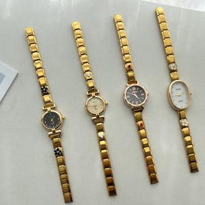 Op de afbeelding: Vijf gouden polshorloges met verschillende wijzerplaatontwerpen. De horloges hebben gouden metalen banden met decoratieve accenten.