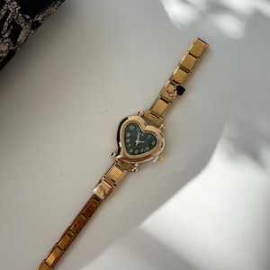 May include: Gold-toned wristwatch with a heart-shaped face and a dark green dial. The watch has a linked band and a small heart charm near the clasp. The watch is on a white surface.