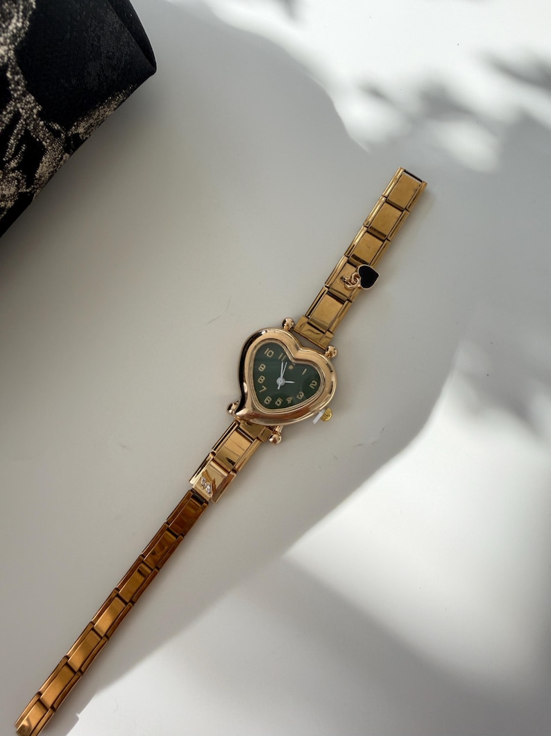 May include: Gold-toned wristwatch with a heart-shaped face and a dark green dial. The watch has a linked band and a small heart charm near the clasp. The watch is on a white surface.