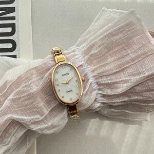 Op de afbeelding: Een ovaalvormig gouden horloge met een witte wijzerplaat en kristallen accenten. De wijzerplaat van het horloge zegt "NAIDU QUARTZ".