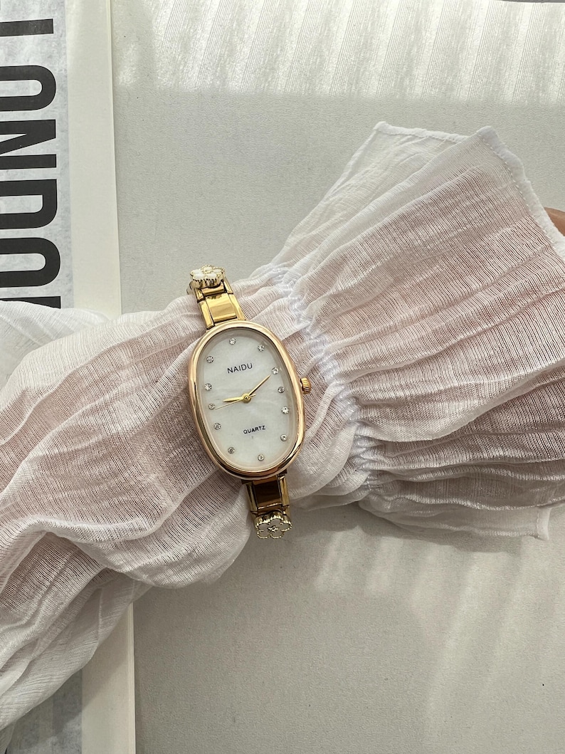Op de afbeelding: Een ovaalvormig gouden horloge met een witte wijzerplaat en kristallen accenten. De wijzerplaat van het horloge zegt "NAIDU QUARTZ".