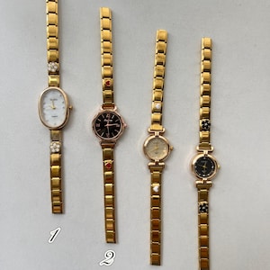 May include: Four gold-toned wristwatches with different face designs and decorative accents. The watches have gold link bands and various face shapes, including oval and round. Each watch features unique embellishments like floral or heart-shaped details.