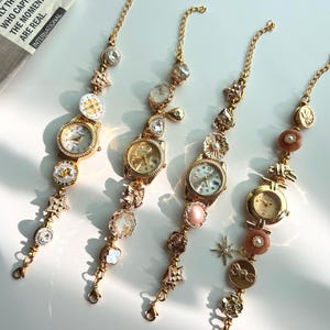 May include: Four gold-toned bracelet watches with decorative charms and watch faces. The watches have various embellishments, including crystals, floral designs, and star-shaped charms. The watch faces have Roman numerals and delicate hands.