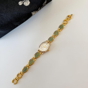 May include: A gold-toned wristwatch with a white oval face and a band featuring green oval-shaped stones. The watch is set against a white surface, with a black floral patterned pouch in the background.