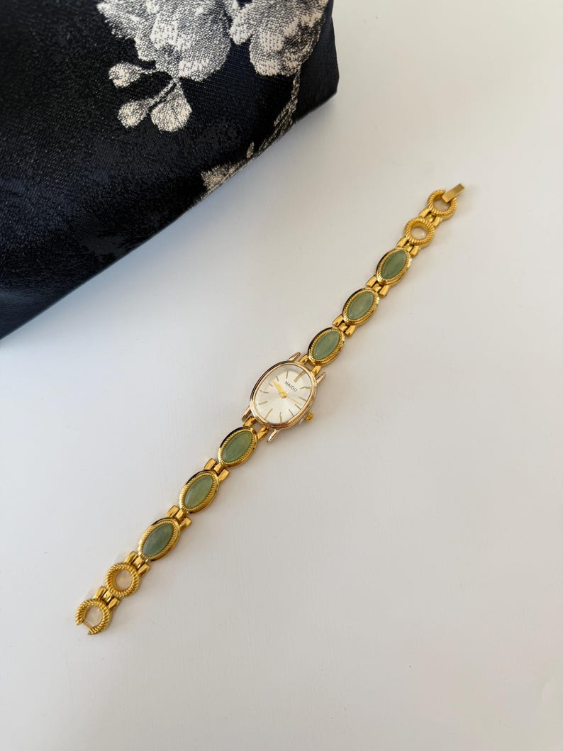 May include: A gold-toned wristwatch with a white oval face and a band featuring green oval-shaped stones. The watch is set against a white surface, with a black floral patterned pouch in the background.