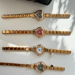 May include: Four gold-toned heart-shaped watches with different colored faces: black, red, white, and green. Each watch has a gold band with decorative elements. One watch has a pearl charm, and another has a spade charm.