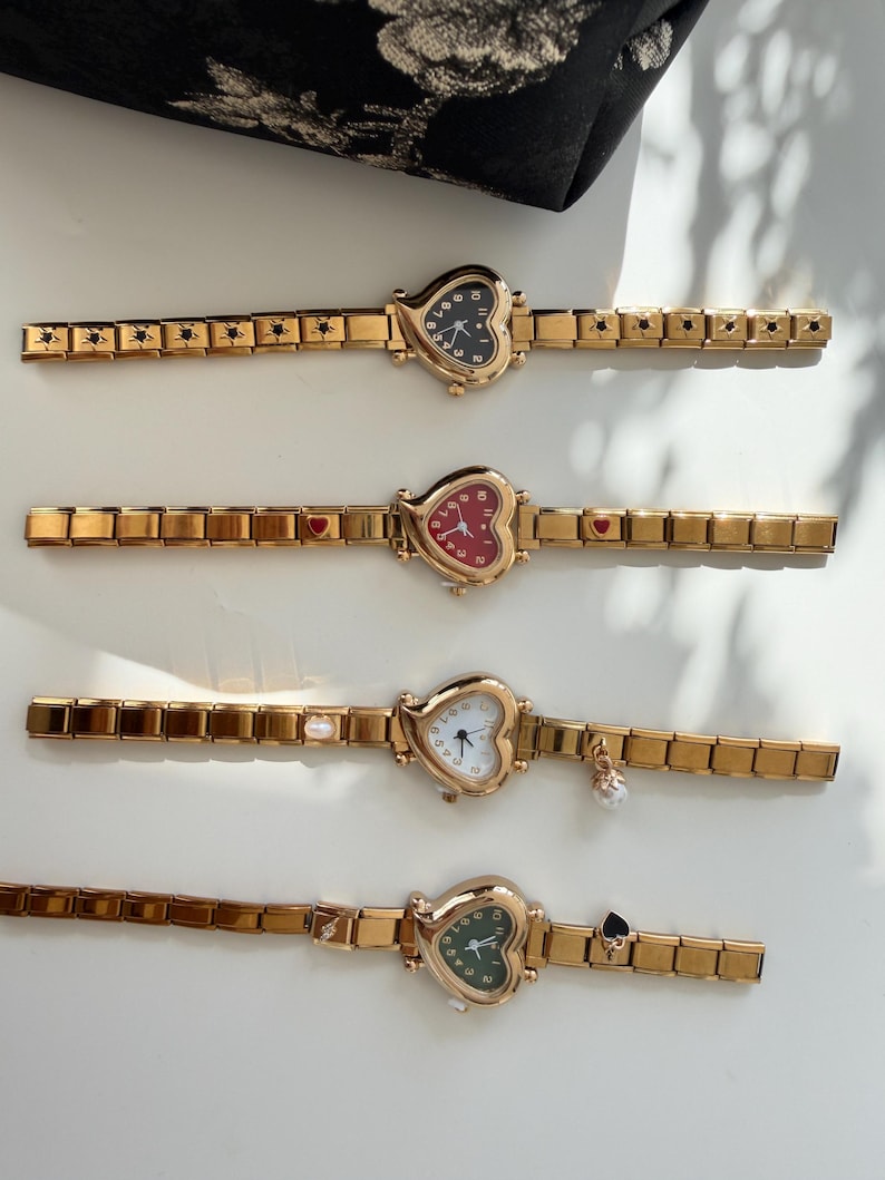 May include: Four gold-toned heart-shaped watches with different colored faces: black, red, white, and green. Each watch has a gold band with decorative elements. One watch has a pearl charm, and another has a spade charm.