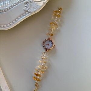 Pu&ograve; includere: Un delicato orologio da polso con cassa in oro rosa e quadrante bianco. Il cinturino &egrave; impreziosito da pietre preziose color ambra e piccole perle bianche. La chiusura &egrave; dorata. L'orologio &egrave; appoggiato su una superficie bianca.