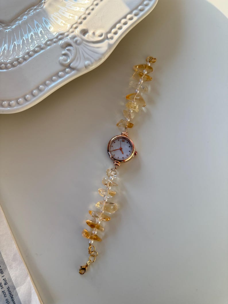 Pu&ograve; includere: Un delicato orologio da polso con cassa in oro rosa e quadrante bianco. Il cinturino &egrave; impreziosito da pietre preziose color ambra e piccole perle bianche. La chiusura &egrave; dorata. L'orologio &egrave; appoggiato su una superficie bianca.