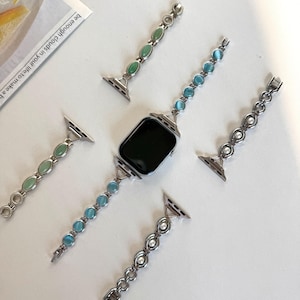 May include: Silver Apple Watch with a black screen, surrounded by four interchangeable bands. The bands feature green, blue, and white decorative stones. The bands are made of silver-colored metal and are designed to be easily swapped out.