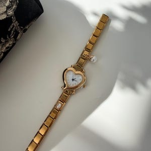 May include: Gold-toned wristwatch with a heart-shaped face and a white dial. The watch features a delicate chain-link band and is adorned with a small pearl charm and a pearl accent on the band. The watch is on a white surface.