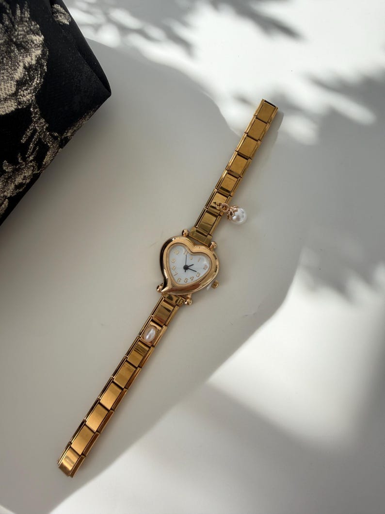 May include: Gold-toned wristwatch with a heart-shaped face and a white dial. The watch features a delicate chain-link band and is adorned with a small pearl charm and a pearl accent on the band. The watch is on a white surface.