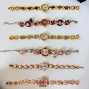 May include: A collection of six vintage-style women's watches. The watches feature gold-toned and silver-toned metal bands with various embellishments, including pearls, gemstones, and floral designs. The watch faces are in various shapes, including round, oval, and heart.