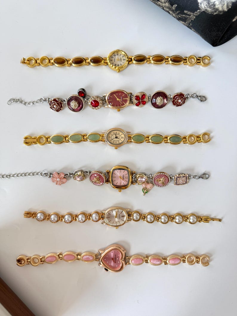 May include: A collection of six vintage-style women's watches. The watches feature gold-toned and silver-toned metal bands with various embellishments, including pearls, gemstones, and floral designs. The watch faces are in various shapes, including round, oval, and heart.