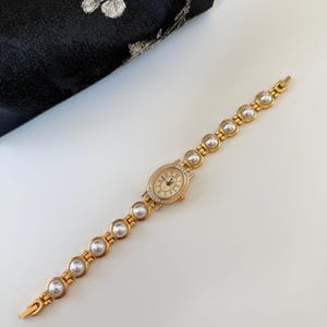May include: A gold-toned wristwatch with a pearl bracelet band. The watch face is oval, with a gold bezel and small crystals. The watch hands are black, and the numbers are gold. The band is made of gold links with large, round pearls.