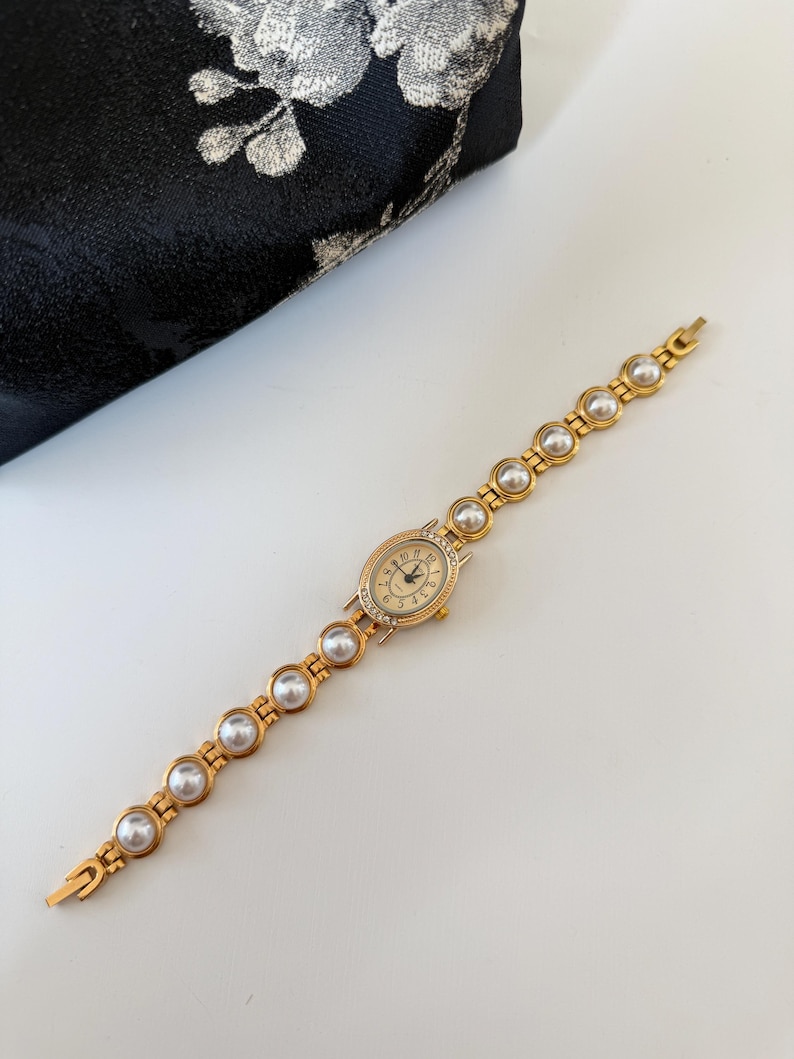 May include: A gold-toned wristwatch with a pearl bracelet band. The watch face is oval, with a gold bezel and small crystals. The watch hands are black, and the numbers are gold. The band is made of gold links with large, round pearls.