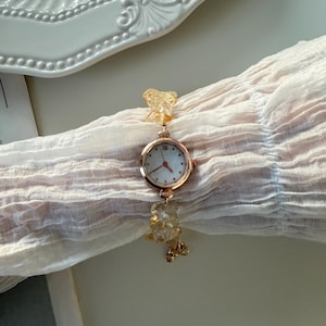 Pu&ograve; includere: Un orologio da polso dorato con quadrante bianco e piccoli indicatori delle ore. Il cinturino &egrave; adornato con perline giallo chiaro di forma irregolare. L'orologio &egrave; esposto su un tessuto trasparente color crema.