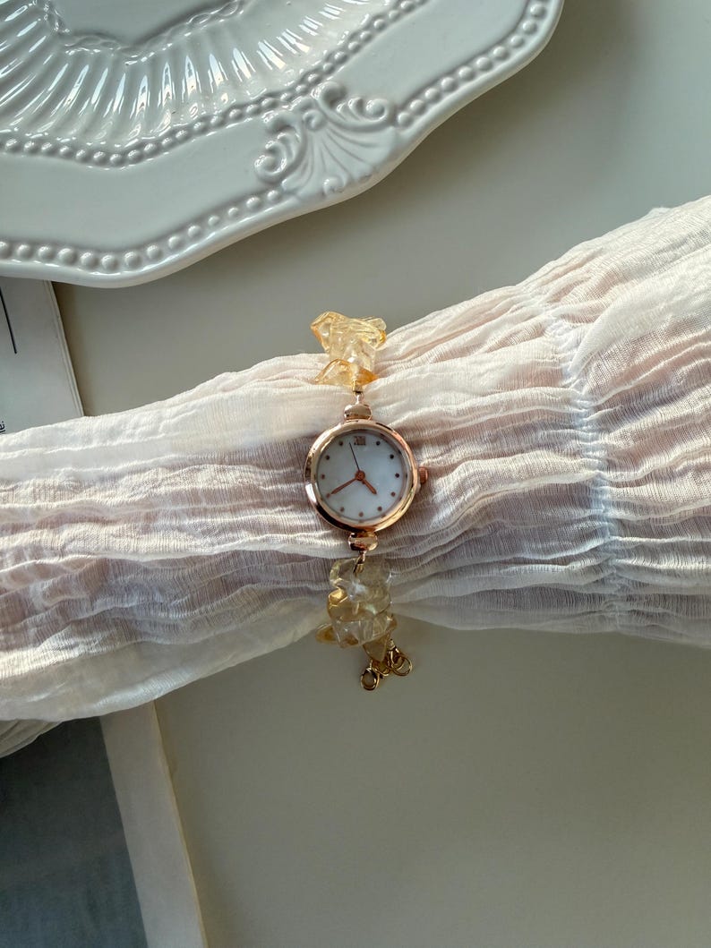 Pu&ograve; includere: Un orologio da polso dorato con quadrante bianco e piccoli indicatori delle ore. Il cinturino &egrave; adornato con perline giallo chiaro di forma irregolare. L'orologio &egrave; esposto su un tessuto trasparente color crema.