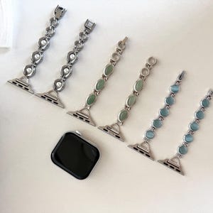 May include: An assortment of watch bands in silver metal with decorative elements. Two bands feature pearls, two have green stones, and two have blue stones. A black smartwatch is also present.