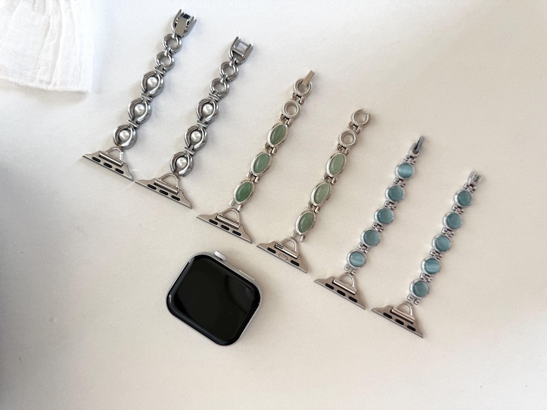 May include: An assortment of watch bands in silver metal with decorative elements. Two bands feature pearls, two have green stones, and two have blue stones. A black smartwatch is also present.
