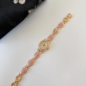 May include: A gold-toned wristwatch with a cream-colored face and pink oval-shaped links. The watch face has small crystals around the edge and black hands. The watch is on a white surface next to a black floral patterned pouch.