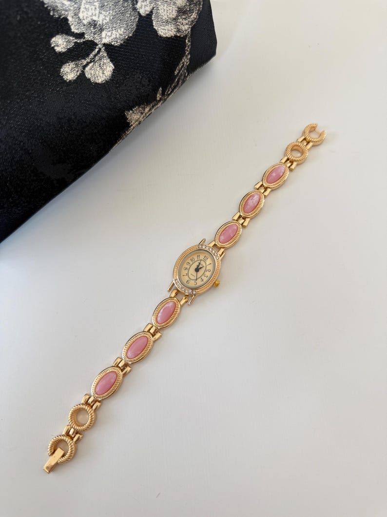 May include: A gold-toned wristwatch with a cream-colored face and pink oval-shaped links. The watch face has small crystals around the edge and black hands. The watch is on a white surface next to a black floral patterned pouch.