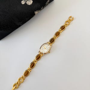 May include: A gold-toned wristwatch with an oval face and a band featuring brown oval-shaped stones. The watch has a white watch face and is set against a white surface. A black clutch with a floral design is in the background.