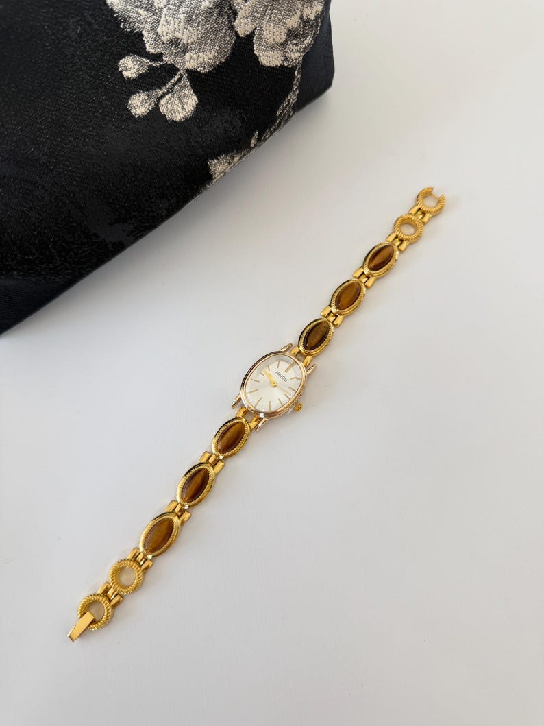 May include: A gold-toned wristwatch with an oval face and a band featuring brown oval-shaped stones. The watch has a white watch face and is set against a white surface. A black clutch with a floral design is in the background.