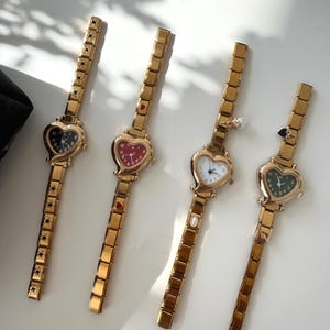 Peut inclure: Quatre montres-bracelets dorées avec des cadrans en forme de cœur. Chaque montre a un cadran de couleur différente : noir, rouge, blanc et vert. Les bracelets de montre sont des maillons rectangulaires. Les montres sont exposées sur une surface blanche.