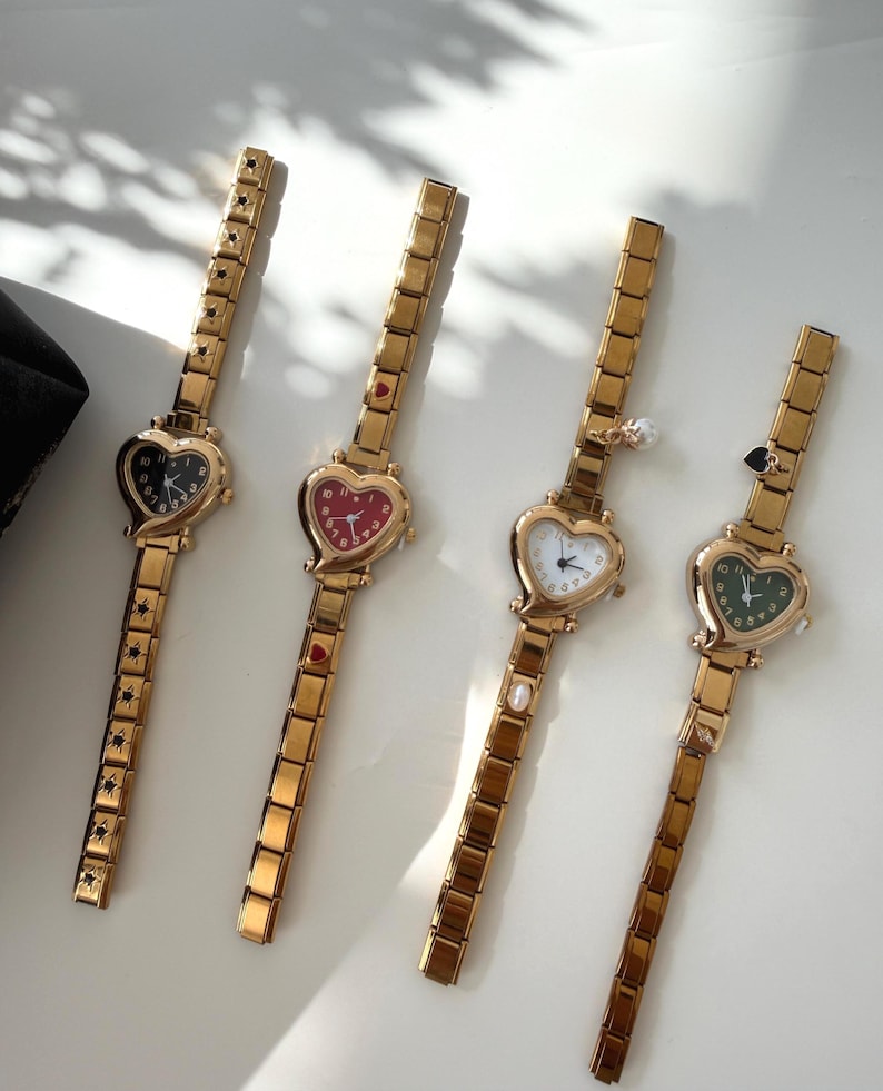 May include: Four gold-toned wristwatches with heart-shaped faces. Each watch has a different colored face: black, red, white, and green. The watch bands are rectangular links. The watches are displayed on a white surface.
