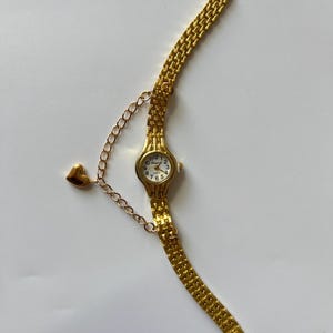Puede incluir: Un reloj de pulsera dorado con un dije en forma de coraz&oacute;n unido a la correa. El reloj tiene una esfera blanca con marcadores de hora en n&uacute;meros romanos negros y un movimiento de cuarzo.