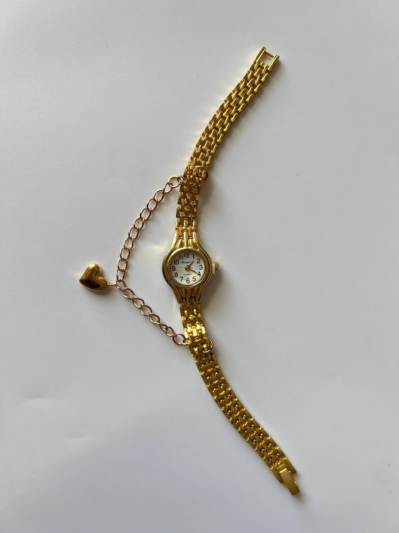 Puede incluir: Un reloj de pulsera dorado con un dije en forma de coraz&oacute;n unido a la correa. El reloj tiene una esfera blanca con marcadores de hora en n&uacute;meros romanos negros y un movimiento de cuarzo.