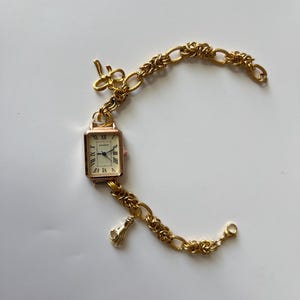 Puede incluir: Un reloj dorado con una esfera cuadrada y n&uacute;meros romanos. El reloj est&aacute; unido a una pulsera de cadena dorada con un dije de llave y un dije de mano.