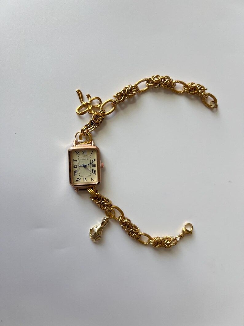 Puede incluir: Un reloj dorado con una esfera cuadrada y n&uacute;meros romanos. El reloj est&aacute; unido a una pulsera de cadena dorada con un dije de llave y un dije de mano.