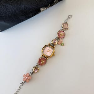 May include: A gold-toned watch with a pink face and decorative charms. The charms include pink flowers, bees, and pink gemstones. The watch is connected to a silver chain. A black pouch with a floral design is in the background.