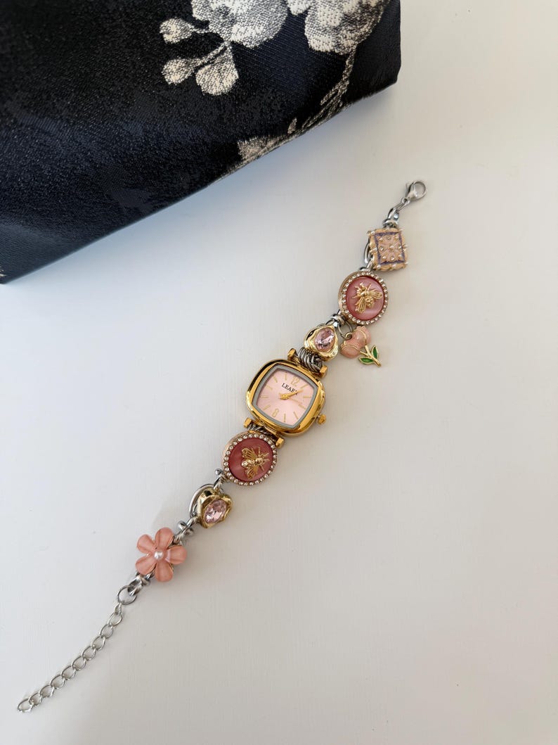May include: A gold-toned watch with a pink face and decorative charms. The charms include pink flowers, bees, and pink gemstones. The watch is connected to a silver chain. A black pouch with a floral design is in the background.