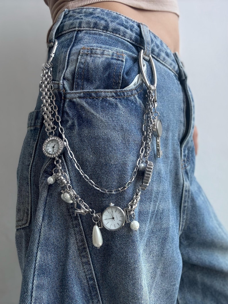 Puede incluir: Accesorio de cadena plateada con m&uacute;ltiples hebras, con peque&ntilde;os relojes redondos, perlas de imitaci&oacute;n y dijes decorativos. La cadena est&aacute; sujeta a unos vaqueros azules.