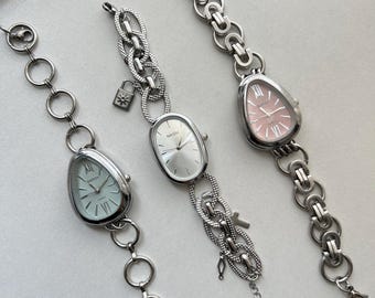 May include: Three silver watches with different colored faces. The watches have unique chain link bands with charms. The watch faces are oval shaped and have Roman numeral markings.