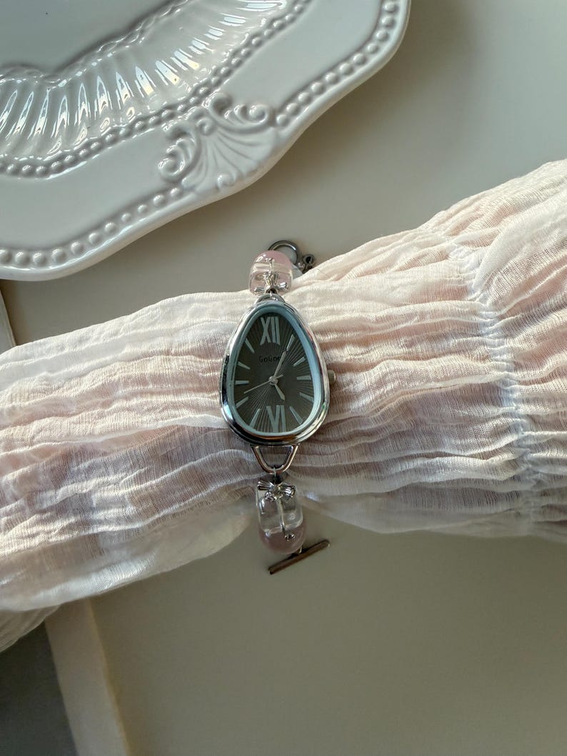 Pu&ograve; includere: Un orologio da polso argentato con quadrante ovale grigio scuro e numeri romani. L'orologio ha un quadrante interno azzurro e dettagli in pietra rosa sul cinturino. L'orologio &egrave; esposto su un tessuto rosa chiaro.