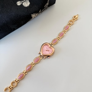 May include: A gold-toned wristwatch with a heart-shaped pink face and a matching pink stone bracelet. The watch features a delicate design with small, round accents and oval-shaped pink stones. The watch is approximately 7 inches long.