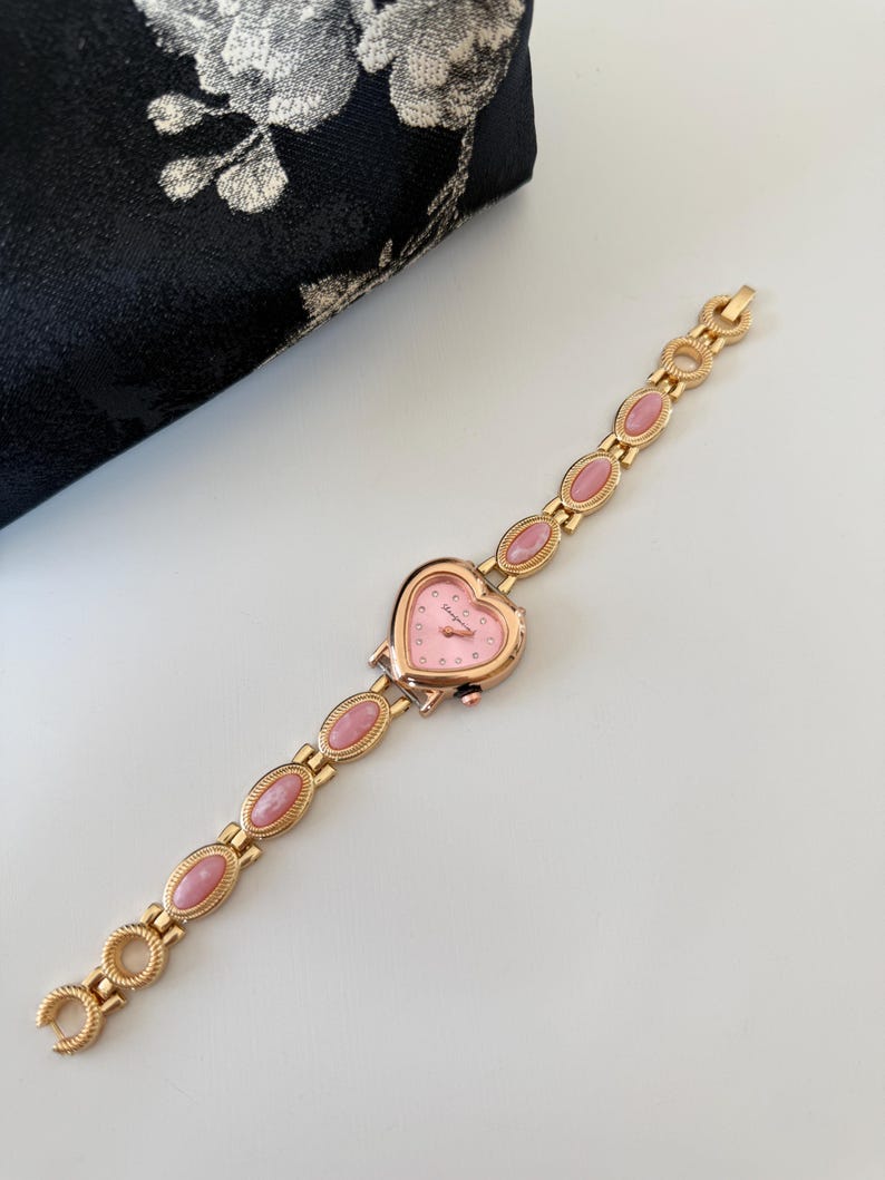 May include: A gold-toned wristwatch with a heart-shaped pink face and a matching pink stone bracelet. The watch features a delicate design with small, round accents and oval-shaped pink stones. The watch is approximately 7 inches long.